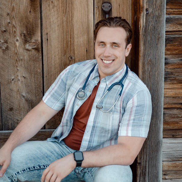 Smiling man in casual attire sitting by a wooden wall.