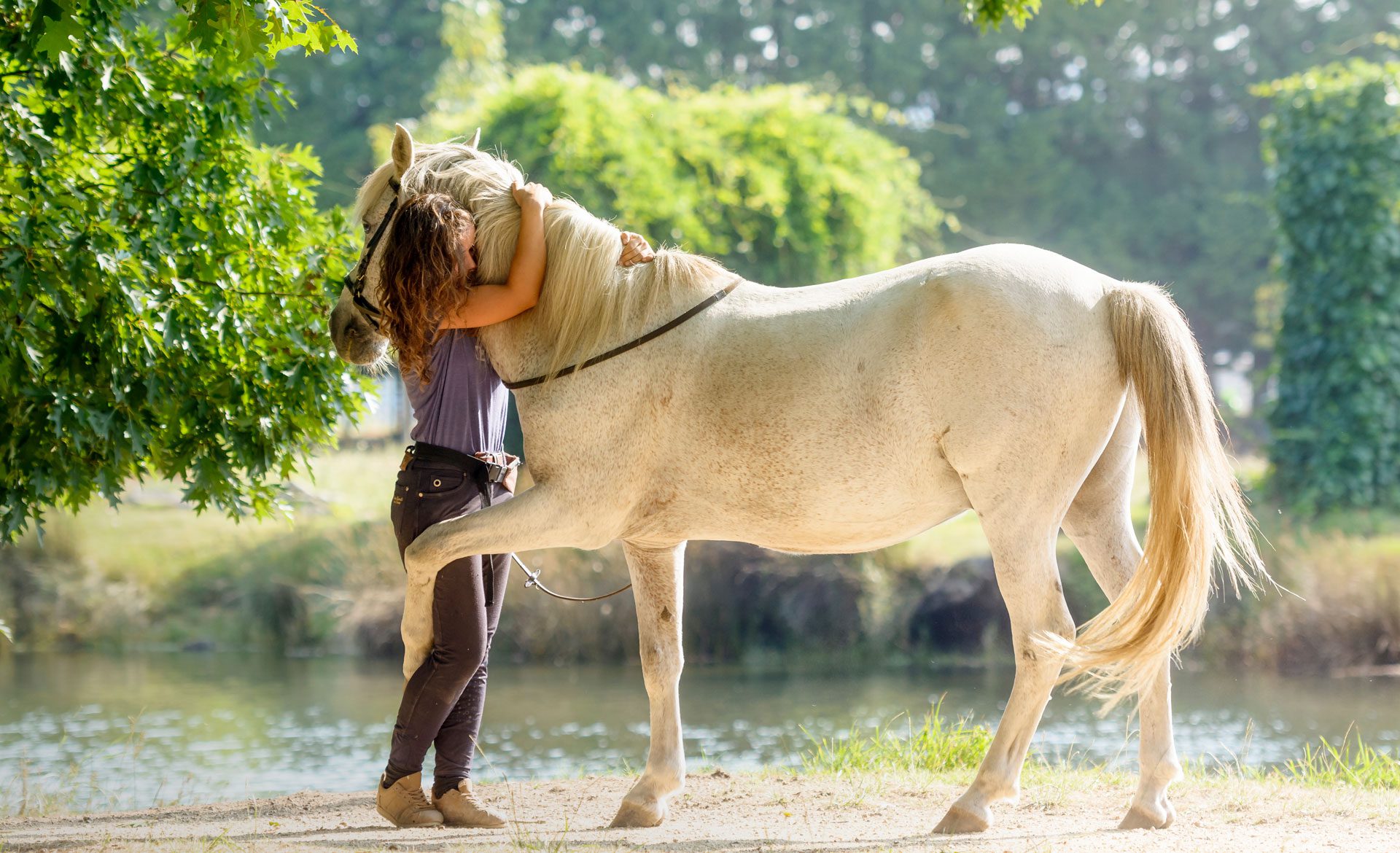 Woman embraces horse outdoors by water.