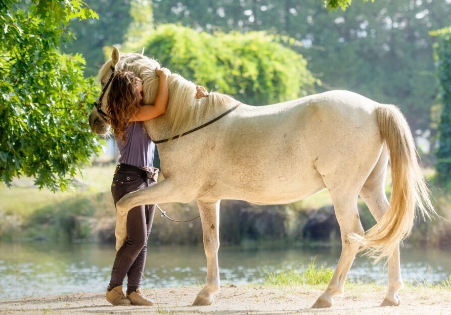 Woman embraces horse outdoors by water.